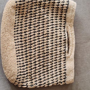 May include: Handmade tote bag with a woven pattern in black and beige. The bag features a rectangular shape with rounded corners and a handle on one side. The woven design creates a textured surface, perfect for carrying essentials.