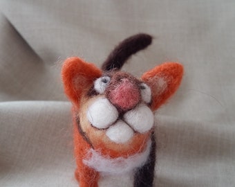 May include: A hand-felted orange and brown cat with white fur on its chest and a pink nose. The cat is standing on a light brown fabric surface.