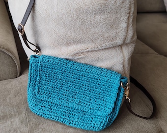 May include: A turquoise crochet crossbody bag with a brown leather strap and gold-colored hardware. The bag is rectangular with a flap closure. The bag is resting on a beige couch with a fluffy white pillow in the background.