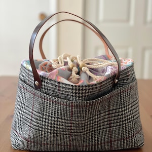 May include: A structured handbag with a black, white, and red plaid exterior. It has brown leather handles and a drawstring closure with wooden beads. The interior lining features a floral pattern.