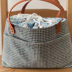 May include: A structured bucket bag featuring a black and white houndstooth pattern on the exterior with two tan leather handles secured by metal rivets. The top opening has a white drawstring closure revealing a blue and yellow floral fabric lining.