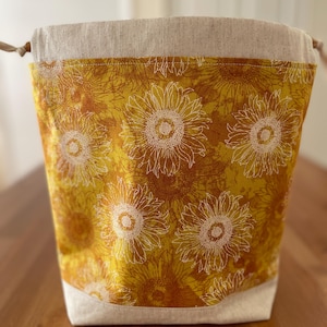 May include: A drawstring bag featuring a vibrant yellow and orange sunflower pattern on the main body, contrasted by a solid light beige fabric at the top and bottom. The bag includes a simple drawstring closure with small wooden beads at the ends.