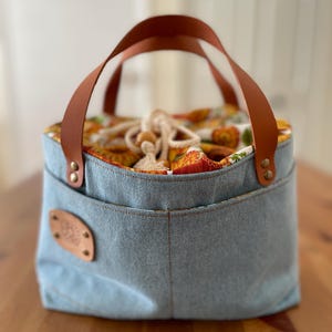 May include: A light blue denim bucket bag featuring two brown leather handles attached with brass rivets. The bag has an exterior pocket and a drawstring closure made of white rope, revealing an interior lining with a colourful sunflower print.