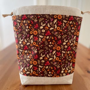 May include: A drawstring bag featuring a dark brown fabric panel with a repeating pattern of red maple leaves, orange sunflowers, acorns, and small mushrooms. The top and bottom sections are made of a light beige, natural-coloured fabric.