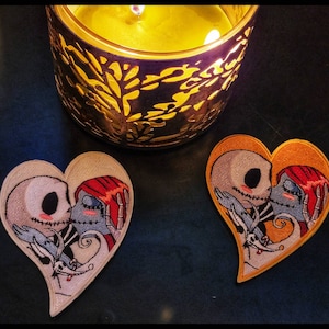 Könnte beinhalten: Zwei herzförmige, gestickte Aufnäher mit Jack und Sally aus The Nightmare Before Christmas beim Küssen. Ein Aufnäher hat einen weißen Hintergrund, der andere einen orangefarbenen Hintergrund. Eine brennende Kerze in einem dekorativen Halter befindet sich im Hintergrund.