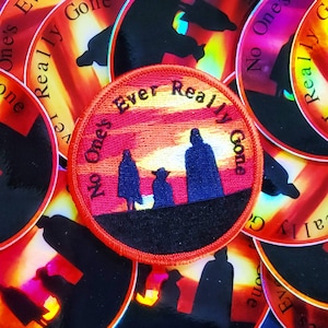 Op de afbeelding: Een ronde, rode geborduurde patch met een zwarte achtergrond. De patch toont een zonsondergangsscene met drie silhouetten tegen een oranje en gele lucht. De tekst "No One's Ever Really Gone" is geschreven in een wit, gebogen lettertype rond de figuren.