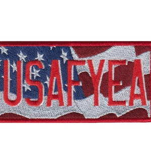 Puede incluir: Parche bordado rojo, blanco y azul con el texto "USA FYEA" sobre un fondo de bandera estadounidense ondeando.