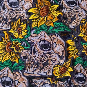 May include: Close-up of embroidered patches featuring a skull with sunflowers. The skull is beige and the sunflowers are yellow with brown centers. The patches are surrounded by green leaves.
