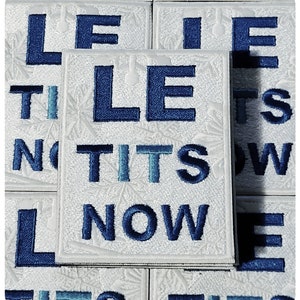Może przedstawiać: Biała naszywka haftowana z niebieskim napisem "Let It Snow" i "Tits Now".
