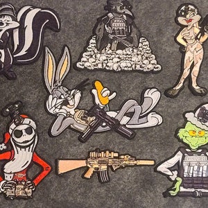 Peut inclure: Un ensemble de neuf patchs brodés représentant divers personnages de dessins animés, dont Bugs Bunny, le Grinch et une moufette. Les patchs sont tous de style militaire, certains comportant des armes et du matériel tactique. Les patchs sont tous sur un fond gris.