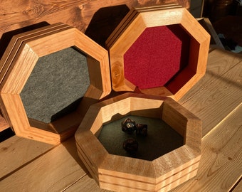 May include: Three wooden octagonal dice trays with felt lining. The trays are in shades of brown and have a natural wood finish. One tray has a red felt lining, one has a gray felt lining, and the other has a green felt lining. There are four black dice in the green tray.