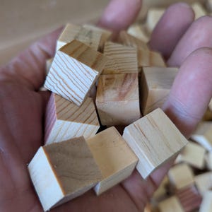 May include: A handful of small, unfinished wooden cubes. The cubes are various shades of light brown and beige, with visible wood grain. Some cubes have a reddish hue. The cubes are likely for crafts or educational purposes.