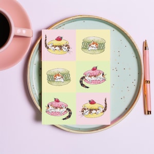 May include: A square art print with six watercolour illustrations of kittens in macarons. Each macaron is a different colour, topped with a strawberry. The print is on a plate with a pink coffee cup and pen.