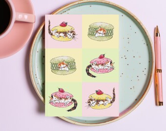Puede incluir: Una impresión artística cuadrada con seis ilustraciones de acuarela de gatitos en macarons coloridos. Cada macaron es de un color diferente, coronado con una fresa. La impresión está sobre un plato con una taza de café y un bolígrafo rosas.