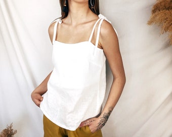 May include: A white sleeveless top featuring a square neckline and thin shoulder straps that tie at the top. The garment is paired with mustard yellow trousers. The top has a straight hemline and a simple, clean design with no visible patterns or prints.