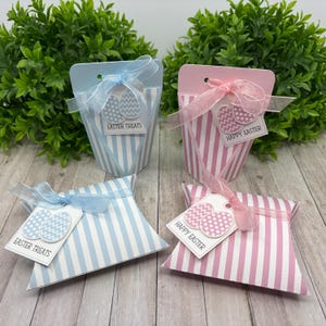 May include: Four Easter-themed gift boxes with striped patterns in blue and pink. Two boxes are upright and two are laying flat. The boxes have tags with the text "Easter Treats" and "Happy Easter" and feature an image of two Easter eggs.