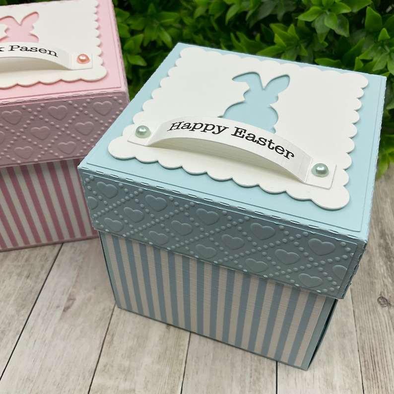 May include: A blue and white striped box with a white lid. The lid has a scalloped edge and a cut-out bunny silhouette. The lid also has a white banner with the text "Happy Easter" on it. The box is decorated with small white hearts.