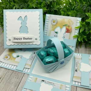 May include: A blue and white Easter explosion box with a bunny silhouette and the words "Happy Easter". The box is open to reveal a blue basket filled with blue and green foil-wrapped chocolate eggs.