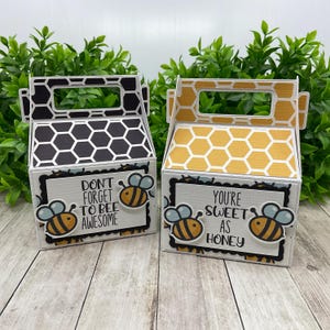 Op de afbeelding: Twee decoratieve geschenkdozen met honingraatpatronen en bijenillustraties. Eén doos heeft de tekst "Don't forget to bee awesome" en de andere zegt "You're sweet as honey". De dozen zijn wit, zwart en geel.
