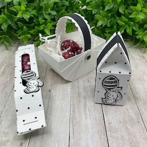 Może przedstawiać: Trzy pudełka na prezenty wielkanocne w biało-czarne kropki. Pudełka mają kształt koszyka, domu i długiego prostokąta. Każde pudełko ma czarno-białego króliczka trzymającego balon z paskowanym jajkiem.