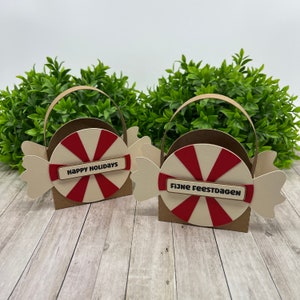 Puede incluir: Dos cestas de regalo con forma de caramelo. Cada cesta tiene una base marrón, un diseño de menta roja y blanca y una envoltura beige. Una dice "Happy Holidays" y la otra "Fijne Feestdagen".