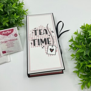 Op de afbeelding: Een zwarte doos met een wit deksel en een rood-zwart lint. Het deksel heeft een rood rasterpatroon met zwarte hartjes en de tekst "TEA TIME" in zwart. Een zwart theezakje-etiket met een rood hartje hangt aan de tekst.