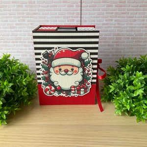 Op de afbeelding: Een rood, wit en zwart gestreepte doos met een illustratie van de kerstman. De doos heeft een rood lint en is versierd met snoepstokken, hulst en andere kerstversiering.
