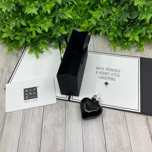 Puede incluir: Una bolsa de regalo negra con una tarjeta de regalo blanca que dice "Rituals Gift Card" y un adorno en forma de corazón negro con el texto "Have yourself a merry little Christmas".