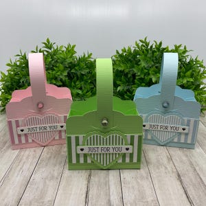 Op de afbeelding: Drie kleine geschenkmanden met handvatten. De manden zijn roze, groen en blauw met witte strepen en een hartvormig ontwerp. Elke mand heeft een label met de tekst "Just for you" met een klein hartje aan beide kanten van de tekst.