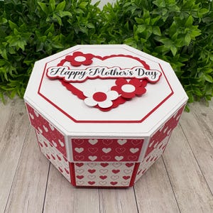 Può includere: Scatola ottagonale bianca con un cuore rosso e fiori rossi e bianchi sul coperchio. Il coperchio ha il testo "Happy Mother's Day" in corsivo nero. La scatola è decorata con un motivo a cuori rossi e bianchi.
