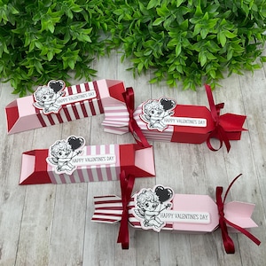 May include: Four red and pink Valentine's Day cracker boxes with a white and black illustration of a cherub holding a heart balloon. Each box has a red ribbon and the text "Happy Valentine's Day".