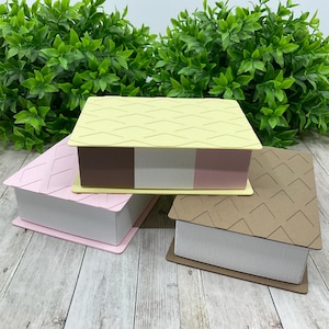 May include: Three decorative boxes with textured lids and white paper sides. The boxes are in pastel colors: pink, yellow, and brown. The lids have a diamond pattern. The boxes are stacked on a wooden surface.