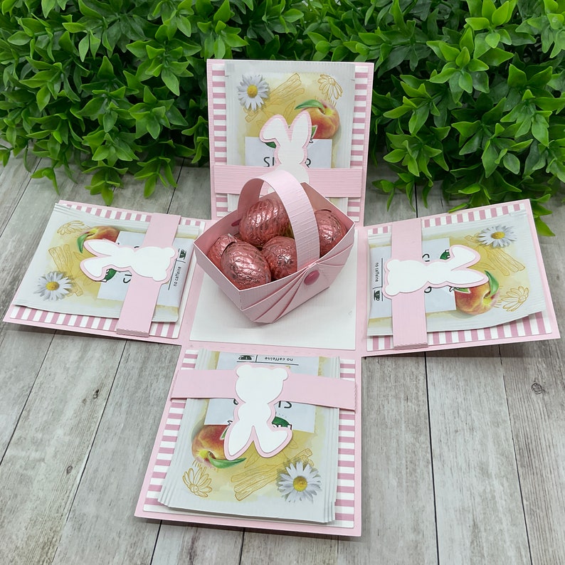 May include: Pink explosion box with a basket of chocolate eggs, bunny cutouts, and a floral design. The box is decorated with pink and white stripes.