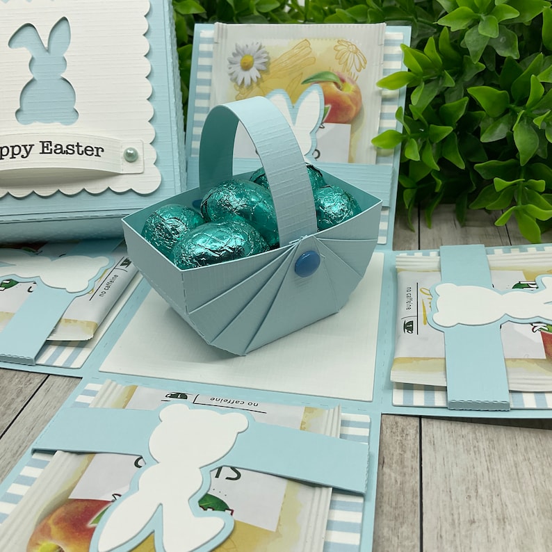 May include: A blue and white Easter-themed explosion box with a bunny rabbit cutout on the front. The box is open to reveal a blue basket filled with chocolate eggs. The box has a white background with blue and white stripes.