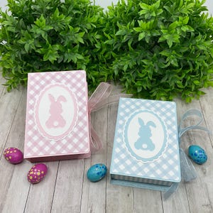 Op de afbeelding: Twee paasgeschenkdozen, één roze en één blauw, met een ruitpatroon en een konijnensilhouet. Elke doos is vastgebonden met een transparant lint. Decoratieve paaseieren liggen rond de dozen.