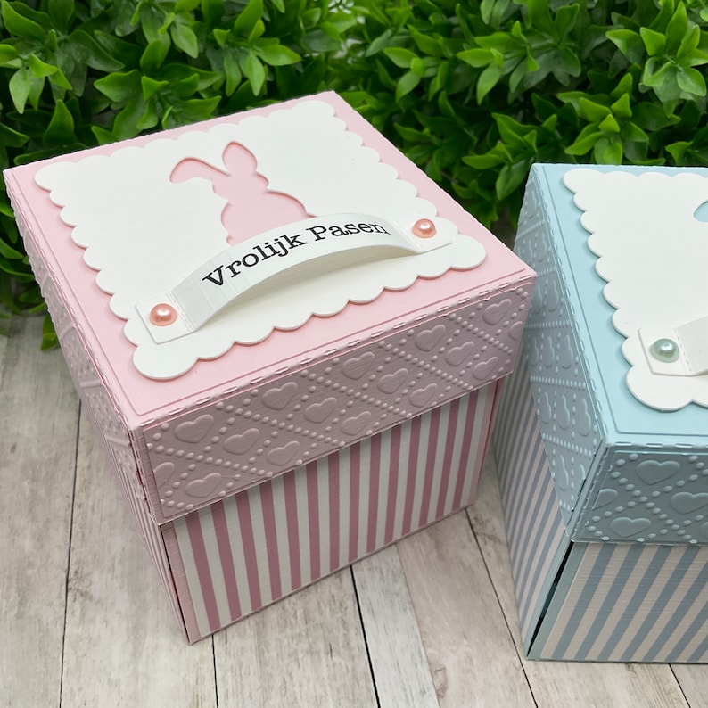 May include: Two small, square gift boxes with a bunny silhouette on the lid. The pink box has a white ribbon with the text "Vrolijk Pasen" and pink pearl accents. The blue box has a white ribbon with pearl accents. Both boxes have a heart pattern and white stripes.