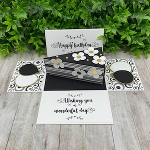 May include: A black and white pop-up card featuring a cake design. The card has a black and white striped cake with white flowers and gold glitter. The card also has a black and white patterned background with the text "Happy birthday" and "Wishing you a wonderful day".