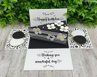 May include: A black and white pop-up card featuring a cake design. The card has a black and white striped cake with white flowers and gold glitter. The card also has a black and white patterned background with the text "Happy birthday" and "Wishing you a wonderful day".