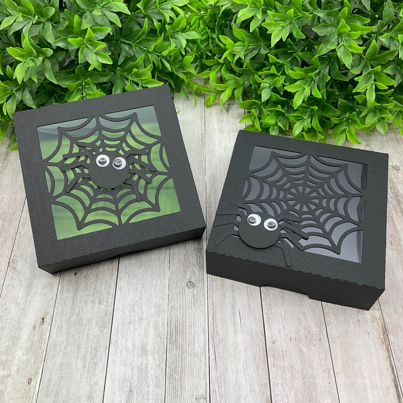 May include: Two black gift boxes with a spider web and spider cut-out design. The boxes have a clear window to show the design.