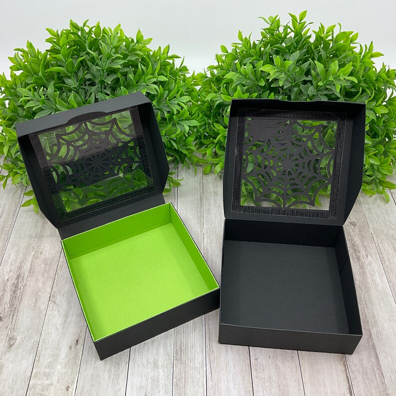 May include: Two black gift boxes with a spiderweb design cut out of the lid. One box has a green interior, the other has a black interior.