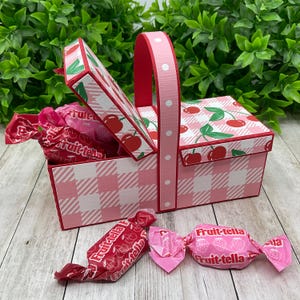 Può includere: Un cestino da picnic rosa e bianco a quadretti con manico rosso e disegno di ciliegie sul coperchio. Il cestino è pieno di caramelle incartate. Due caramelle sono davanti al cestino.