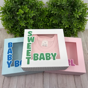 Op de afbeelding: Drie cadeaudozen met uitgesneden ramen, elk met een andere kleur en ontwerp. De bovenste doos is wit met groene letters die "SWEET BABY" zeggen en een roze shirt erin. De middelste doos is lichtblauw met blauwe letters die "BABY BOY" zeggen. De onderste doos is roze met roze letters die "GIRL" zeggen.
