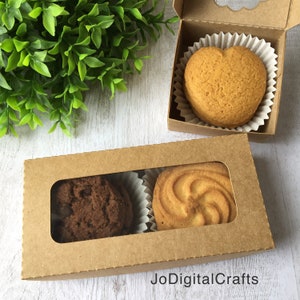 Op de afbeelding: Twee doorzichtige dozen met ramen en een bruine papieren buitenkant. Eén doos bevat twee koekjes, één chocolade en één swirl. De andere doos bevat een hartvormig koekje.