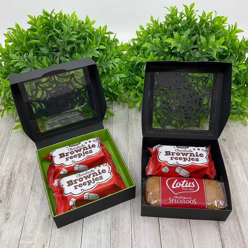 May include: Two black gift boxes with clear lids featuring a spiderweb design. The boxes are filled with chocolate brownie bars and a package of Lotus Speculoos cookies.