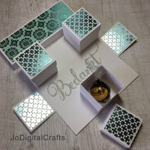 Może przedstawiać: Białe pudełko z wysuwanym wiekiem ozdobione papierem w kolorze turkusowym i srebrnym. Pudełko jest otwarte i zawiera małą czekoladkę. Pudełko jest oznaczone napisem "Bedankt" w srebrnym brokacie.