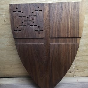 May include: A wooden shield with a dark brown finish. The shield has a cross design and a series of cut-out crosses on the upper left quadrant. The shield is a decorative item.