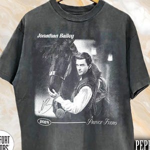 May include: Pepper-colored t-shirt featuring a black and white image of Jonathan Bailey with a horse. The text "Jonathan Bailey," "Prince Fyero," and "2024" are also visible. The shirt is from Comfort Colors.