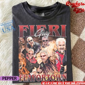 May include: A charcoal gray t-shirt featuring a graphic print of Guy Fieri in various poses against a background of flames. The text FIERI GUY appears in large red letters at the top, and the word FLAVORTOWN is printed in red at the bottom.