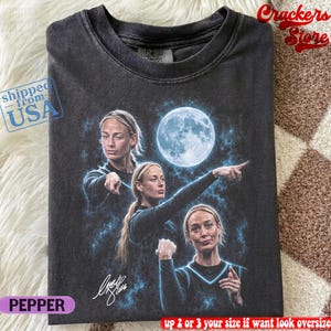 Puede incluir: Una camiseta de cuello redondo gris carbón con un estampado gráfico de una persona en tres poses diferentes sobre un fondo cósmico azul con una luna llena. La camiseta incluye una firma y una etiqueta de la marca Comfort Colors en el cuello.