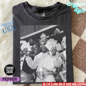 Könnte beinhalten: Pfefferfarbenes Comfort Colors T-Shirt mit einem Schwarz-Weiß-Bild eines Boxers, der Bargeld und einen Meisterschaftsgürtel hält. Das Bild enthält den Text "TRUMP PLAZA". Das Shirt ist mit "shipped from USA" gekennzeichnet.